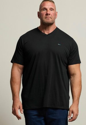 Muskeløs mand med kort hår og skæg iført en ensfarvet sort V-hals T-shirt og mørke jeans, stående mod en neutral baggrund.