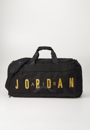 Bolso de deporte negro con la letra "JORDAN" en dorado. Presenta un destacado logo negro de Jumpman, dobles asas superiores y una correa ajustable.