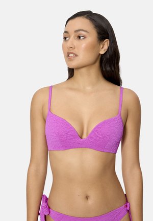 Mujer con cabello largo y oscuro, vistiendo un bikini texturizado morado con la parte superior y la parte inferior a juego, esta última con lazos laterales, mirando ligeramente hacia la izquierda sobre un fondo liso.