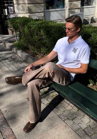 Homme portant un polo blanc et un pantalon beige assis en tailleur sur un banc vert dans une zone urbaine ensoleillée avec des arbustes.