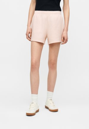 Personne portant un short rose clair à taille élastique, des chaussettes blanches et des baskets crème avec des semelles marron, debout devant un fond blanc uni.