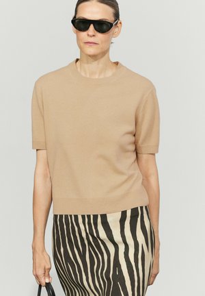 Kvinde iført sorte solbriller, beige kortærmet sweater og sort-hvid stribet nederdel, der holder en sort håndtaske.