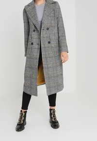 Lång rutig kappa i grått och svart, med dubbelknäppning, breda slag och gul foder, kombinerad med svarta leggings och stövlar.