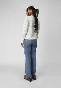 White lace long-sleeve top with puff sleeves, paired with blue straight-leg jeans. The model stands with braided hair, facing away from the camera.