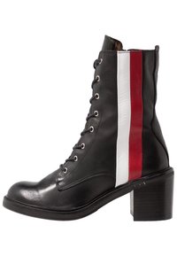 Bottine en cuir noir avec un talon épais, présentant des rayures verticales blanches et rouges sur les côtés et des œillets en métal pour les lacets.