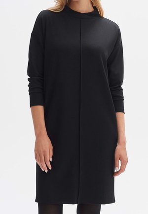 Woman wearing a long-sleeve, loose-fitting black dress with a high neckline and center seam detail, standing against a plain background.
