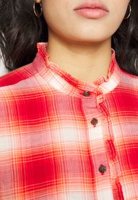 Kobieta w czerwono-pomarańczowej koszuli w kratę z guzikami i falbaniastym, wysokim kołnierzem, nosząca złote kolczyki w kształcie kółek, z długimi, ciemnymi włosami widocznymi.