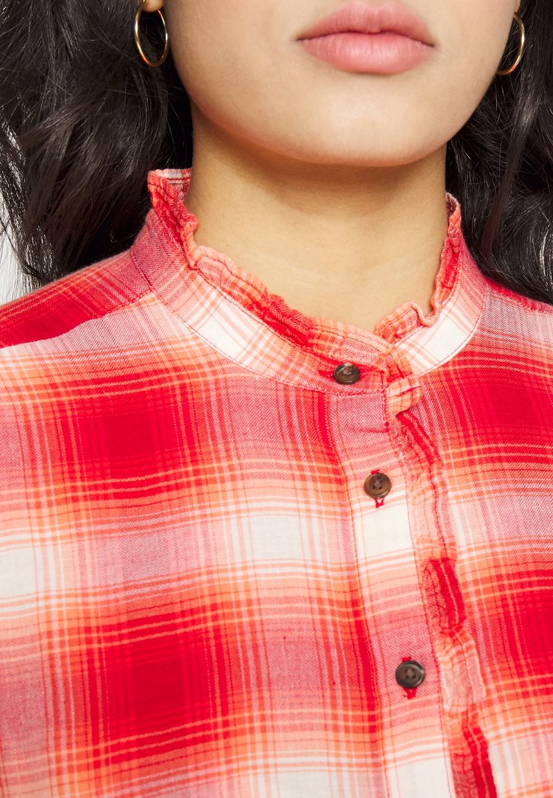 Kobieta w czerwono-pomarańczowej koszuli w kratę z guzikami i falbaniastym, wysokim kołnierzem, nosząca złote kolczyki w kształcie kółek, z długimi, ciemnymi włosami widocznymi.