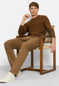 Brauner Strickpullover, khakifarbene Hose und helle Sneakers. Sitzend auf einem Holzstuhl mit geflochtenem Sitz. Neutrale Hintergrundfarbe, Fokus auf die Details der Kleidung.