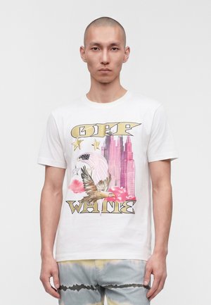 Jeune homme portant un t-shirt blanc avec un graphisme d'aigle, des étoiles, des gratte-ciels roses et le texte « OFF WHITE ».