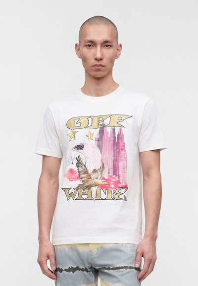 Jeune homme portant un t-shirt blanc avec un graphisme d'aigle, des étoiles, des gratte-ciels roses et le texte « OFF WHITE ».
