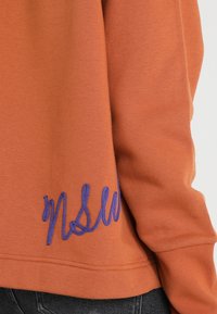 Orange sweatshirt i texturerat tyg, med en lila broderad "NSW"-logotyp på ärmen. Enkel design med en avslappnad passform.