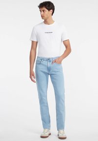 Svijetloplave ravne traperice s prednjim džepovima, uparene s bijelom majicom koja ima crni logotip "GUESS JEANS", nosi se s bež i bijelim tenisicama.
