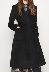 Svart kappa gjord av mjukt tyg, med omslagdesign, knytbälte, två framfickor och slagkrage.