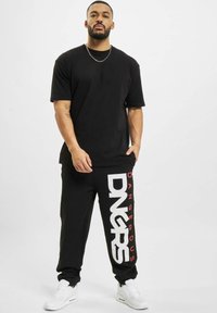 T-shirt nera e pantaloni joggers neri con grafica "DNGRS" bianca e rossa. Il modello indossa sneakers bianche e una collana in argento. Design semplice.