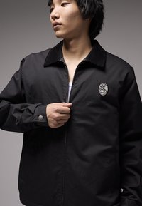 Μαύρο μπουφάν από λείο ύφασμα, με γιακά από κοτλέ, φερμουάρ μπροστά και στρογγυλή επιγραφή στο στήθος.