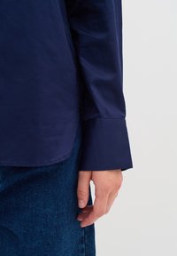 Camicia bottone blu navy con una texture liscia, caratterizzata da un colletto classico e maniche lunghe, abbinata a jeans blu scuro.