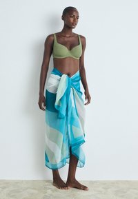 Bralette verde y una bufanda azul con estampado atada a la cintura. La bufanda es transparente con formas geométricas en azul y blanco. Descalza sobre un suelo claro.
