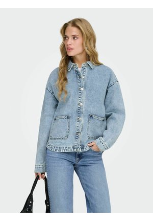 Naine kannab helesinist teksajopet ja teksapükse, hoiab musta käekotti, vaatab küljele helel ühevärvilisel taustal.