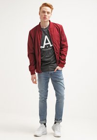 Rdeča bomber jakna preko sivega T-shirt-a z grafiko, v kombinaciji s svetlo modrimi kavbojkami in belimi supergami, stoji ob nevtralni ozadju.