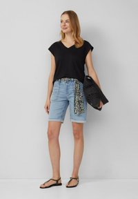 Vrouw in een zwarte mouwloze V-hals top, lichte denim Bermuda shorts met een patroonriem, houdt een zwarte geweven tas vast, draagt zwarte sandalen, op een neutrale achtergrond.