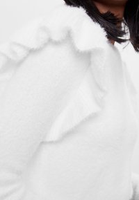 White sweater featuring soft, fuzzy texture and ruffled accents along the shoulders. Prominent lightweight and cozy fabric design.