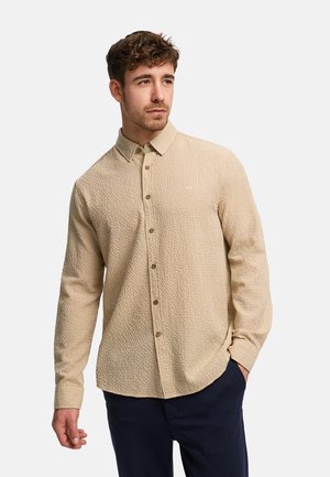 Mand iført beige struktureret skjorte med knapper og mørke bukser, stående med den ene hånd i lommen mod en ensfarvet baggrund.