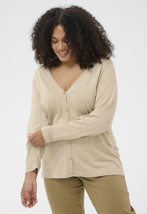 Usmievavá žena s kučeravými vlasmi, oblečená v béžovom zapínanom kardigáne a kaki nohaviciach, upravujúca rukáv na bielom pozadí.