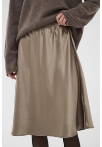 Jupe beige en satin avec une taille élastique, longueur genou et texture lisse. Associée à un pull marron, révélant une partie du vêtement.