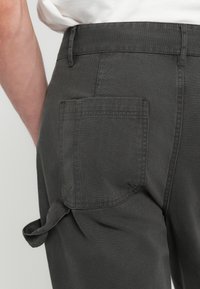Graue Baumwollhose mit einer Gesäßtasche im Quadratdesign, dekorativen Nähten und einer Stoffschlaufe an der Seite.