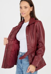 Kvinde iført bordeaux skindjakke, der er åben for at vise det glatte for, hvid ribbet top og blå højtaljede jeans mod en ensfarvet baggrund.