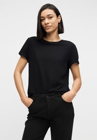 Γυναίκα με μαύρα μαλλιά που φοράει μαύρο t-shirt με ρολαρισμένα μανίκια και μαύρο παντελόνι, στέκεται με τα χέρια στις τσέπες μπροστά σε ένα απλό φωτεινό φόντο.