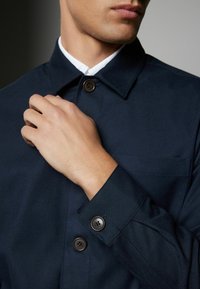 Navy blue shirt jacket with a spread collar, two brown buttons on the cuff, and a front pocket, featuring a smooth, textured fabric.