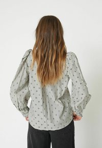 Striped blouse in light grey with puffy sleeves and elastic cuffs, featuring black dots. Loose fit, back view, paired with dark pants.