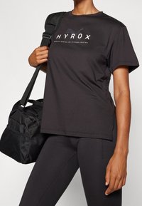 Camiseta atlética negra con mangas cortas y cuello redondo, con "HYROX" impreso en letras blancas en el frente. Acompañada de una bolsa de gimnasio negra.