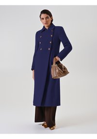 Cappotto doppiopetto blu navy con bottoni dorati, maniche lunghe e una forma strutturata. Il modello tiene una borsa in pelle marrone con dettagli in catena.