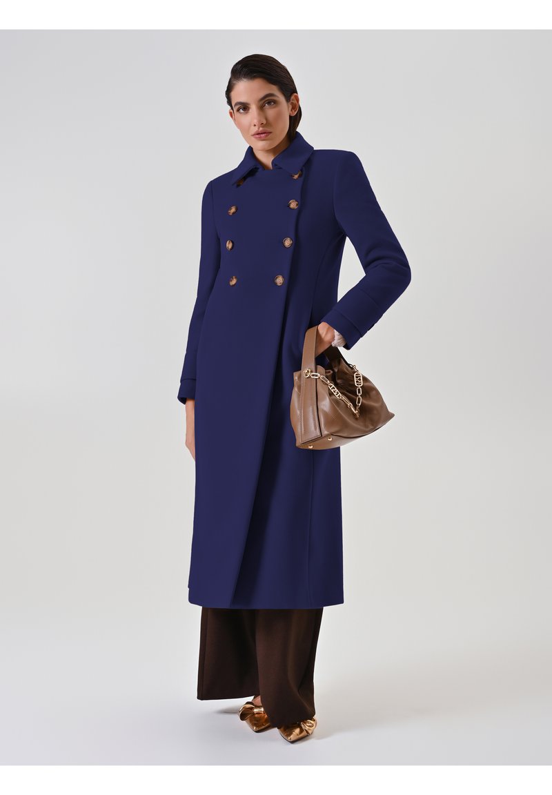 Cappotto doppiopetto blu navy con bottoni dorati, maniche lunghe e una forma strutturata. Il modello tiene una borsa in pelle marrone con dettagli in catena.