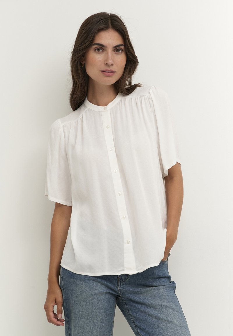White short-sleeve blouse with a round neckline, button closure, and pleated detailing on shoulders. Fabric appears lightweight and slightly textured.