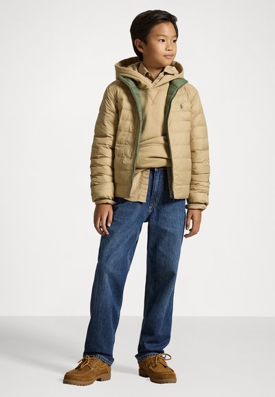 Light beige puffer jacket with green accents, layered over a beige hoodie. Dark blue jeans and brown suede shoes complete the outfit.