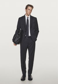 Mörkgrå kostym i strukturerat tyg, slim-fit design, vit skjorta, enfärgat mörkt slips och svarta läderskor. Bär en svart väska.