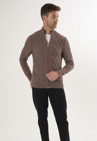 Bruine gebreide zip-up cardigan met een diamant kabelpatroon, geribde manchetten en zoom, gedragen over een witte shirt, gecombineerd met zwarte broek.