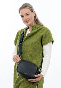 Borsa a tracolla in pelle nera con finitura texture, forma ovale, tracolla regolabile e branding sottile. Indossata su un capo di abbigliamento verde con texture.