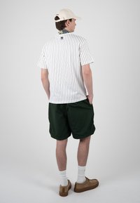 Jeune homme portant un T-shirt blanc à fines rayures, un short vert foncé, une casquette beige, des chaussettes blanches et des chaussures marron, debout les mains dans les poches.