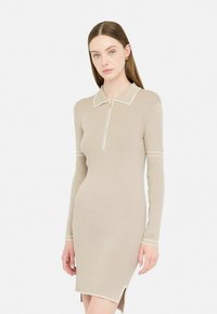 Abito beige a coste con maniche lunghe, colletto e una cerniera a metà. Caratterizzato da rifiniture bianche e fessure laterali per un tocco di dettaglio e texture.