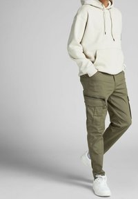 Beige hoodie med framficka, tillsammans med gröna cargobyxor med dragkedjefickor. Bärs med vita sneakers mot en neutral bakgrund.