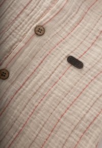 Tejido crema ligero con rayas verticales rojas, que presenta botones de madera redondos y una pequeña etiqueta negra con texto en relieve.