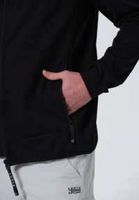 Schwarze Softshelljacke mit Reißverschlusstaschen und verstellbaren Bündchen, die ein schlankes Design und eine leichte Textur aufweist. Logo auf der Hose sichtbar.