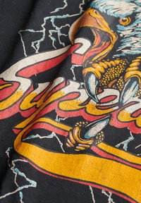 T-shirt nera in cotone con un vivace design grafico di un'aquila con artigli, circondata da testi audaci in rosso e giallo e motivi di fulmini.