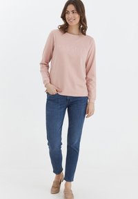 Sudadera rosa claro con la palabra "PASIÓN" bordada en un tipo de letra ligeramente elevada, combinada con jeans de mezclilla oscura y mocasines beige.