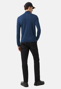 Mann in einem marineblauen Pullover, schwarzen Jeans, schwarzen Schuhen und einer schwarzen Mütze, der mit den Händen in den Taschen stehend nach hinten schaut.